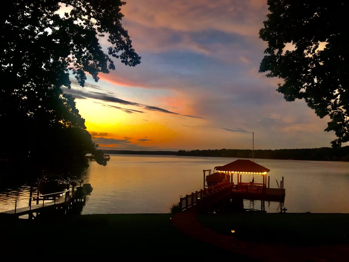 A serene waterfront scene captures a docked gazebo illuminated by soft lights against a vibrant sunset. The calm lake reflects hues of orange and pink, flanked by lush green trees. The overall setting evokes tranquility, inviting relaxation by the water.