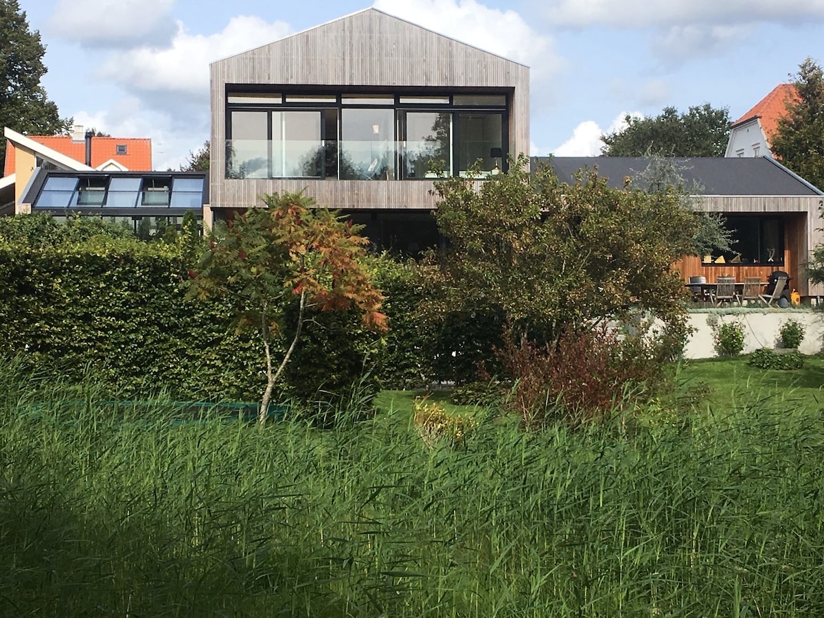 A modern architectural design is showcased with wooden cladding and expansive glass windows. The building is surrounded by lush greenery and a well-maintained garden, offering a serene environment. Outdoor seating is visible on the terrace, providing a comfortable spot for relaxation.