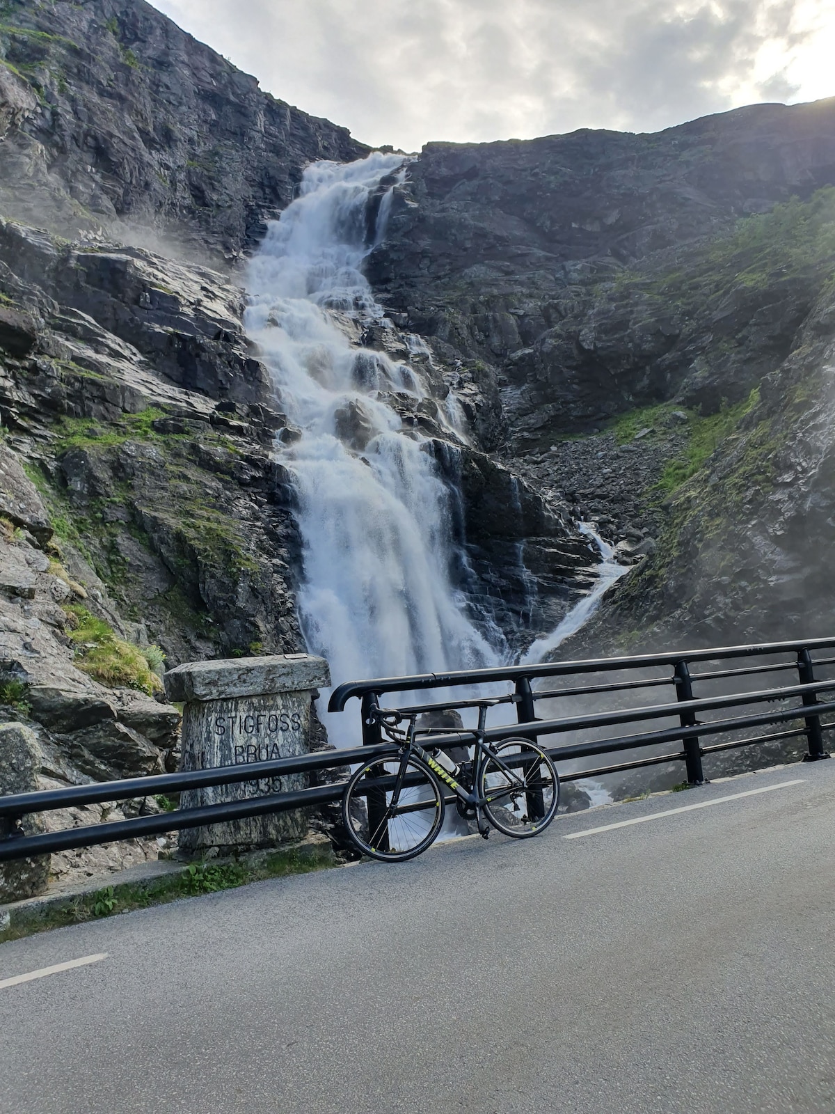 A road runs alongside a waterfall cascading down rocky cliffs. The scene is enhanced by the mist rising from the rushing water. A bicycle is parked on the road, highlighting the accessibility of the area for outdoor activities. Lush greenery frames the landscape.