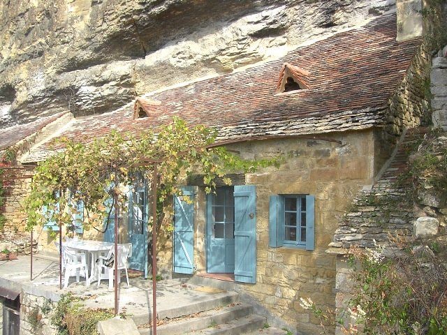 A charming stone house is nestled against a rock face, showcasing a rustic sloped roof and blue shutters. A covered terrace is visible, featuring a white table and chairs. Natural greenery is seen climbing the stone structure, enhancing the serene outdoor setting.