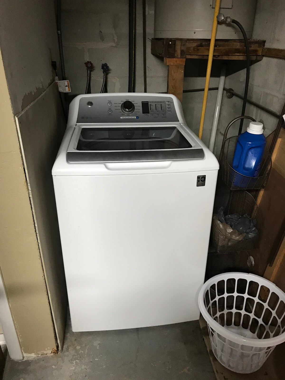 A washer is positioned against the wall in a dimly lit laundry area. A large white laundry basket is placed nearby on the floor. Pipes and various items are visible on the shelving above, contributing to a functional laundry space.
