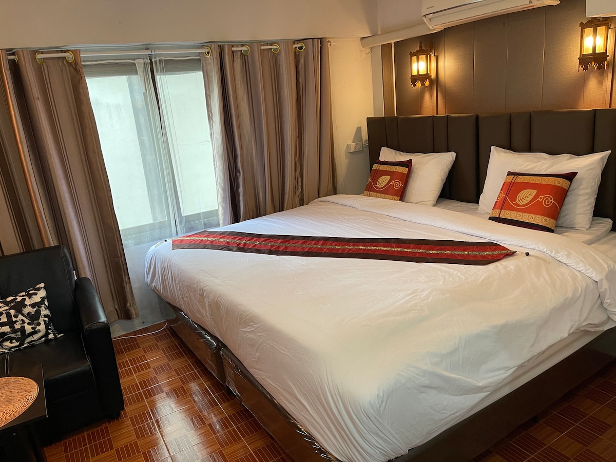 A spacious room features a king-size bed with comfortable linens and decorative pillows. Soft lighting from wall sconces complements the large windows dressed with light curtains. A cozy seating area with a small table is visible, adding to the room's functionality.
