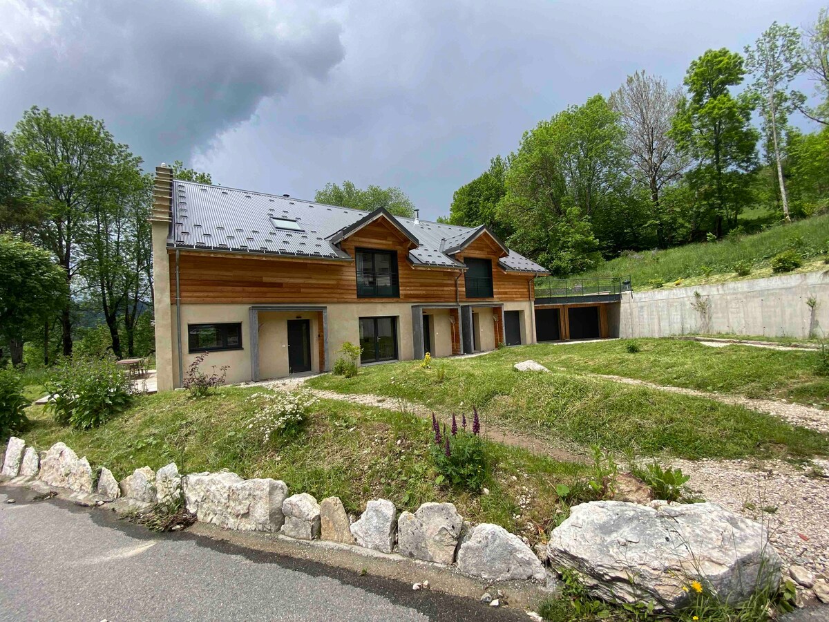 A modern building featuring a combination of wood and stone finishes, set amidst lush greenery. The structure includes multiple entrances, and a sloped garden area with neatly arranged pathways. Trees and wildflowers enhance the natural surroundings, providing a serene atmosphere.