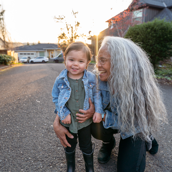 A smiling person with long wavy silver hair crouches next to a smiling toddler in rubber boots. Sunlight streams behind.