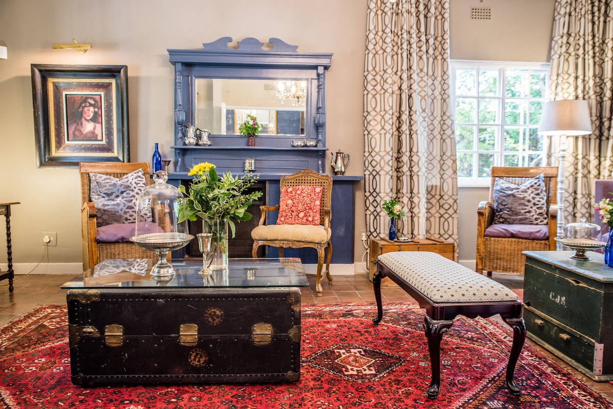 A communal sitting room features comfortable seating with two armchairs and a decorative bench. A vintage coffee table sits atop a patterned area rug, while a mirror and artwork adorn the walls. Natural light filters through the large window, highlighting the inviting decor.