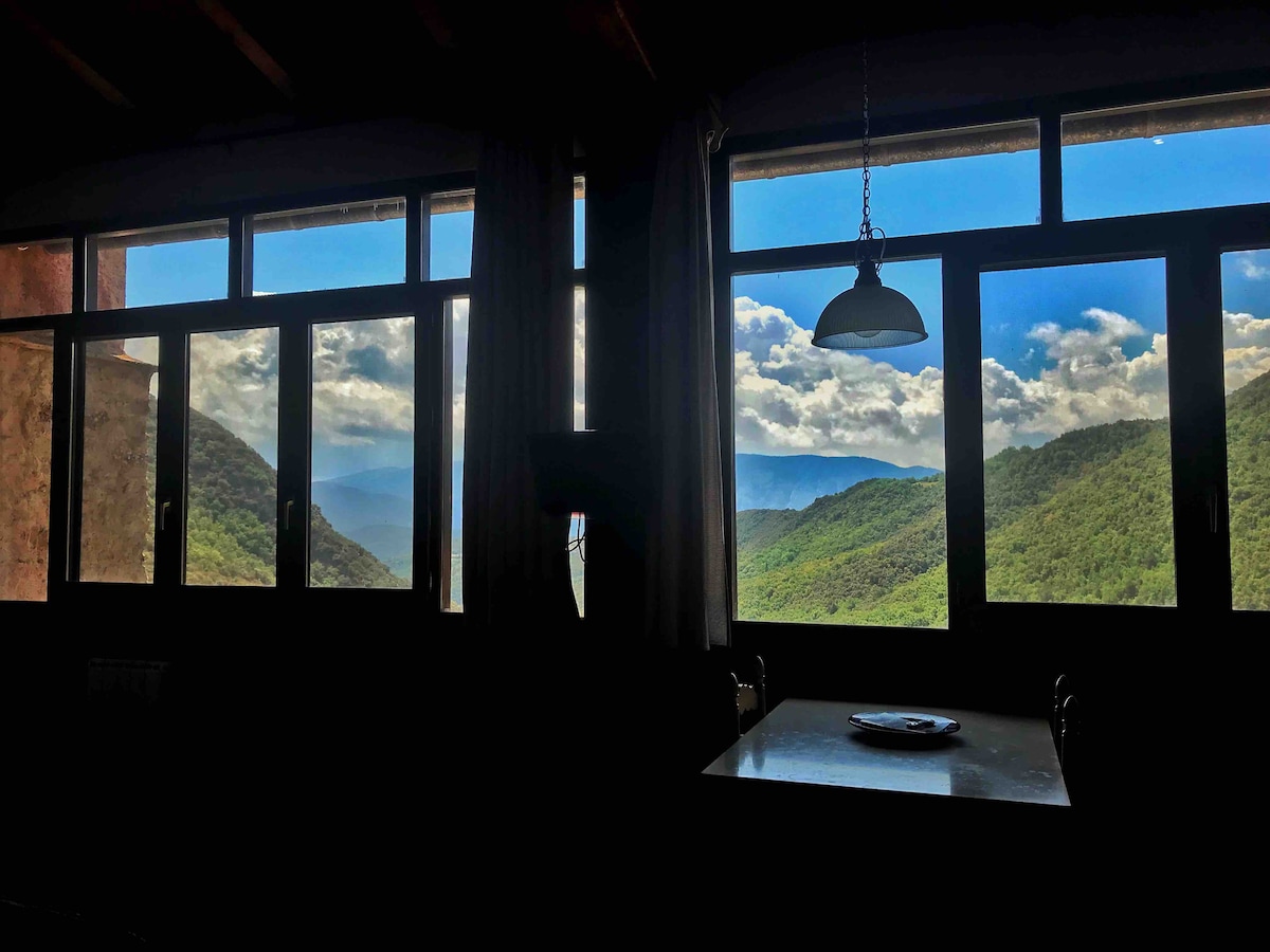 Large windows frame sweeping views of green mountains and a blue sky dotted with clouds, allowing natural light to fill the open space. A dining table is positioned near the windows, creating an inviting area for meals while enjoying the surrounding nature.