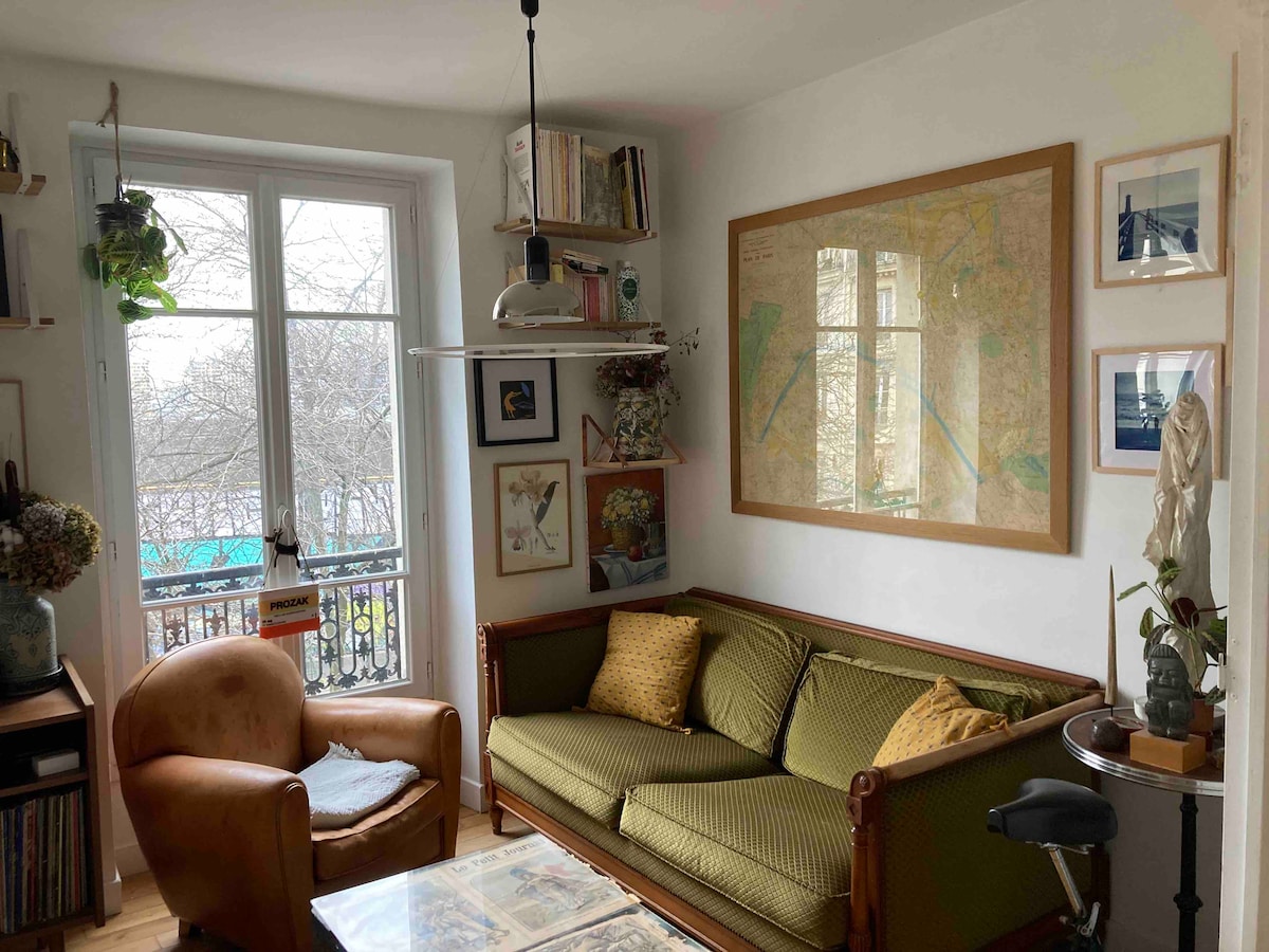 The living area includes a vintage green couch adorned with yellow cushions, paired with a comforting armchair. A map hangs on the wall above a small table. Natural light filters through a large window, which offers views of the outdoors, enhancing the inviting space.