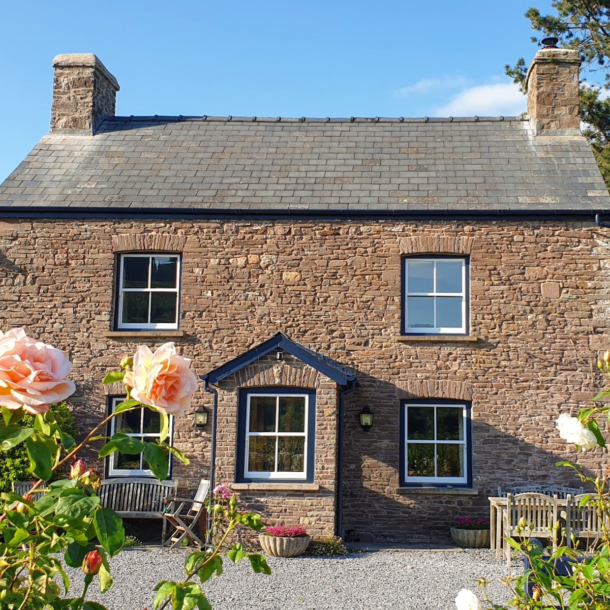 The cozy cottage features a rugged stone façade, complemented by large windows that bring in natural light. A flowerbed with blooming roses frames the space, while a gravel pathway leads to a seating area, enhancing the tranquil outdoor setting.