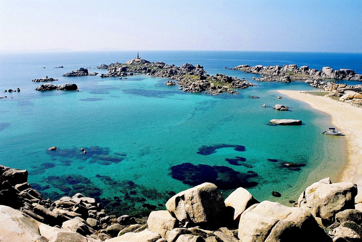 A panoramic view showcases rocky coastal formations and clear turquoise waters. The shoreline features patches of sandy beach, while a distant lighthouse stands atop a rocky outcrop. The sky is clear, creating a serene atmosphere that highlights the area's natural beauty.