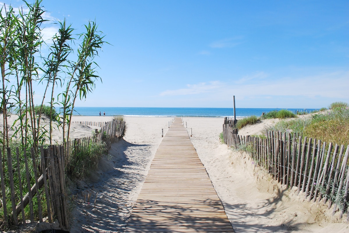 A wooden pathway leads from a sandy area toward the beach, flanked by vegetation and rustic fencing. The beach extends into the distance, with calm ocean waves visible under a clear blue sky.