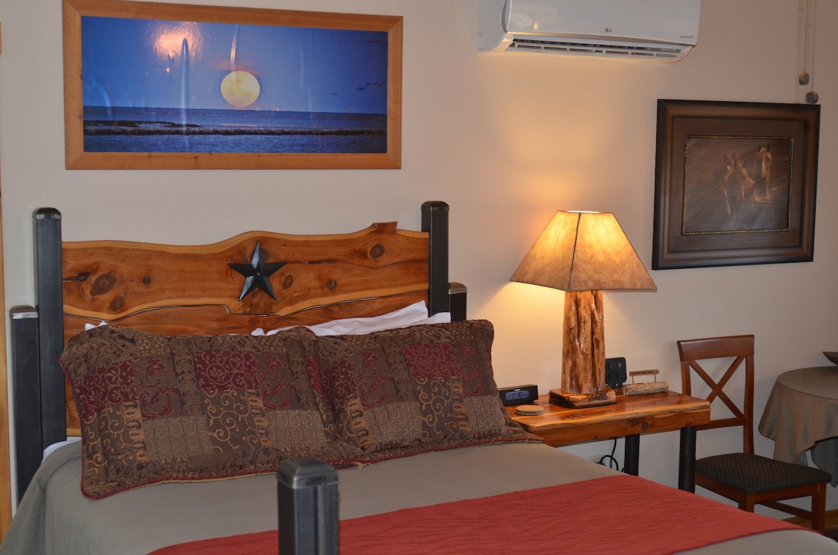A spacious bed is centered against a wooden headboard featuring a star design. A decorative lamp sits on a wooden bedside table, illuminating the area. A piece of art depicting a moonlit sky hangs above the bed, enhancing the room's rustic charm.