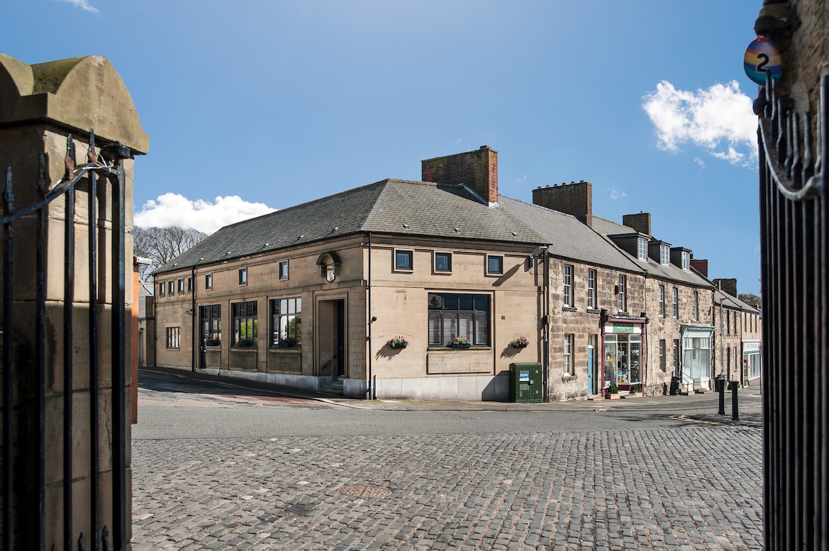 The exterior of Bank View Holiday Cottage is showcased, revealing a charming stone building with large windows reflecting the bright sky. The cobblestone street in front adds character, while surrounding structures hint at a friendly village atmosphere.