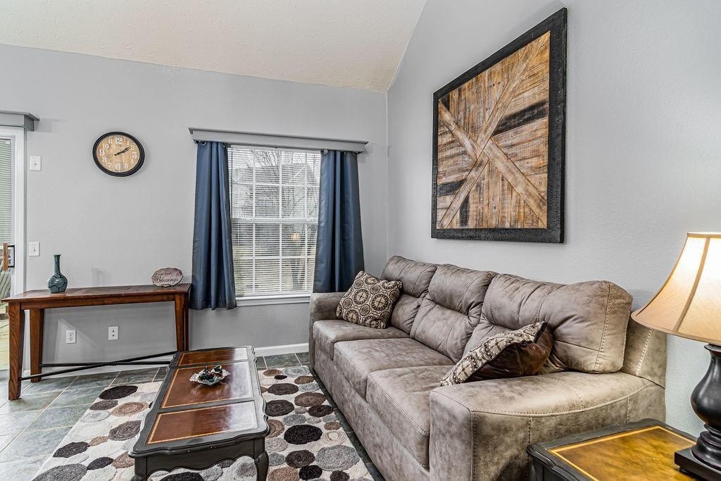 A spacious living area features a large sectional sofa adorned with patterned cushions. A rectangular coffee table sits in front of the sofa, while a decorative piece is displayed on an adjacent wooden table. Natural light enters through a sizable window, framed by dark curtains.