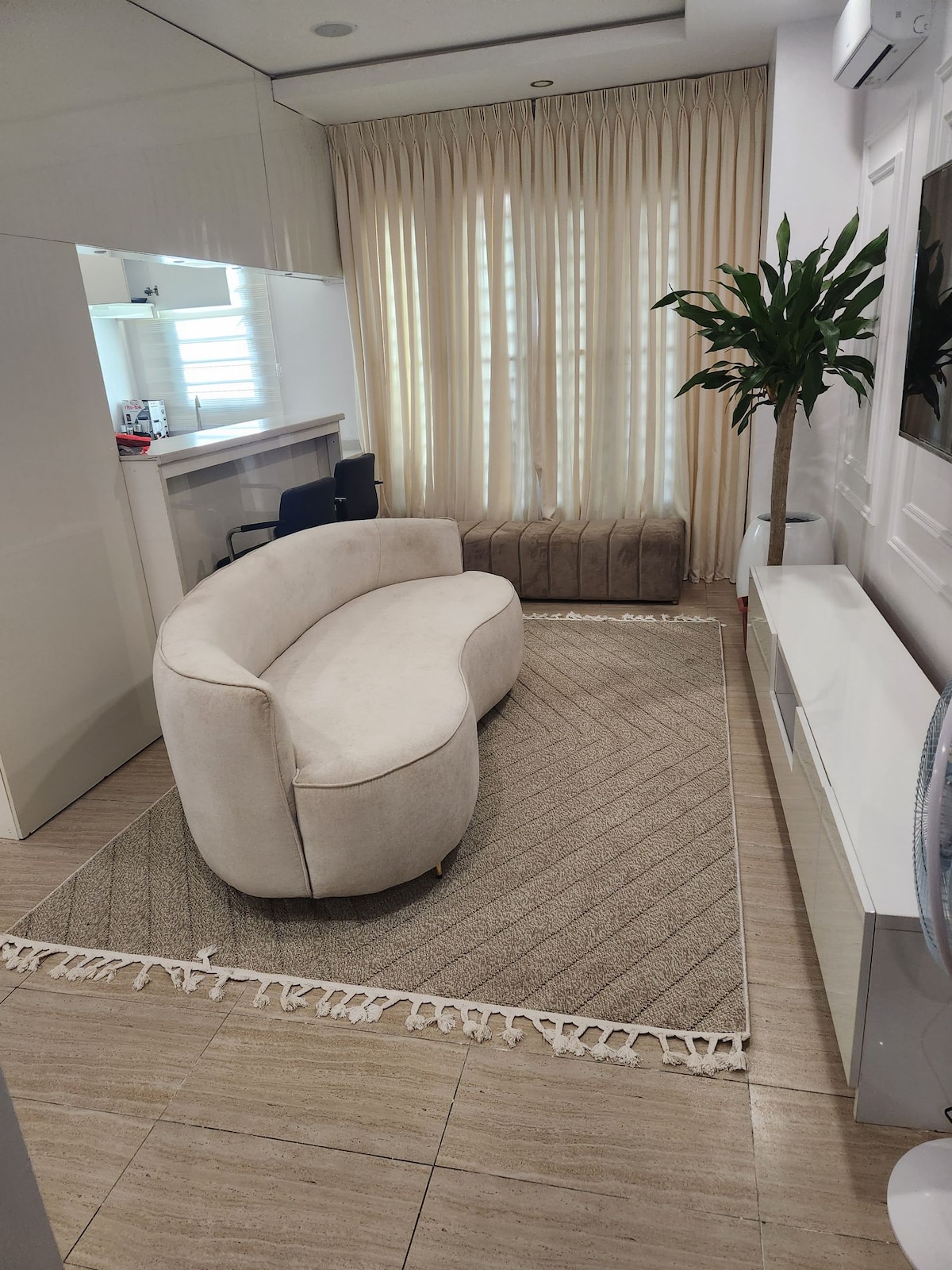 A softly curved sofa is situated on a textured area rug, with a plant adding a touch of greenery. Light filters through floor-to-ceiling sheer curtains, enhancing the bright and airy feel of the living area, which features sleek modern furnishings.