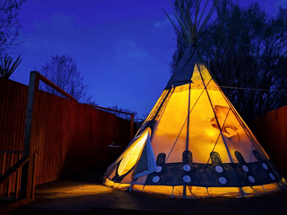A spacious family tipi is illuminated from within against a twilight sky. The structure features a circular design with a distinctive pointed top and is surrounded by wooden fences. The warm light emanating from inside highlights the tipi's unique texture and shapes.