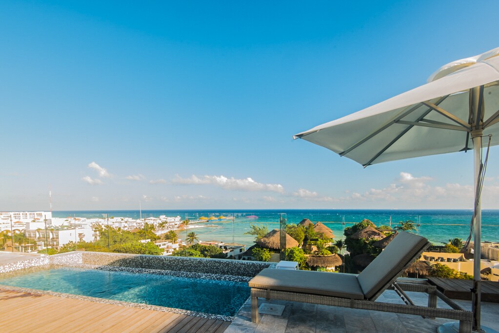 A rooftop area features a lounge chair beside an infinity pool, with sweeping views of the ocean and nearby coastline. A large umbrella provides shade, creating a relaxing ambiance under the clear blue sky.