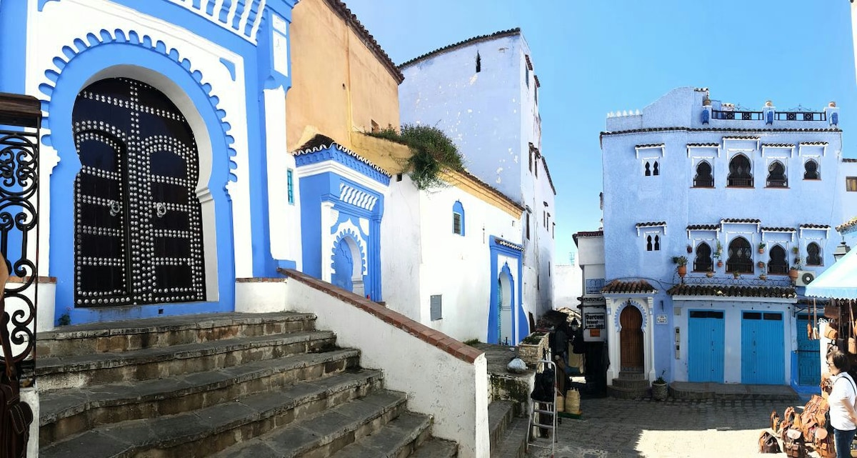 The image captures a charming street scene in Chefchaouen, featuring traditional blue and white buildings. A decorative doorway with intricate patterns draws attention, while stone steps lead down to the street, where crafts and local life can be observed. Clear skies frame the vibrant architecture.