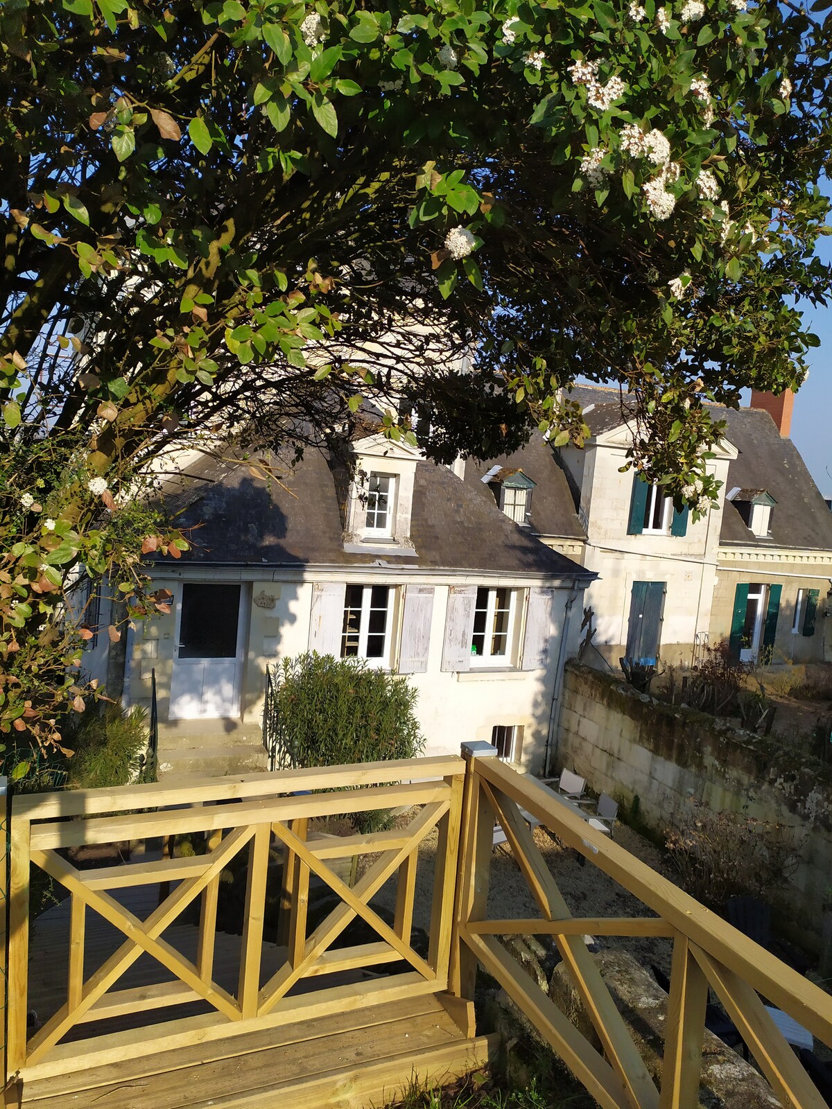 A charming gîte is visible, featuring traditional architecture and exterior windows framed in blue. A wooden deck with a railing is presented in the foreground, surrounded by greenery, while the structure is partially shaded by a blossoming tree.