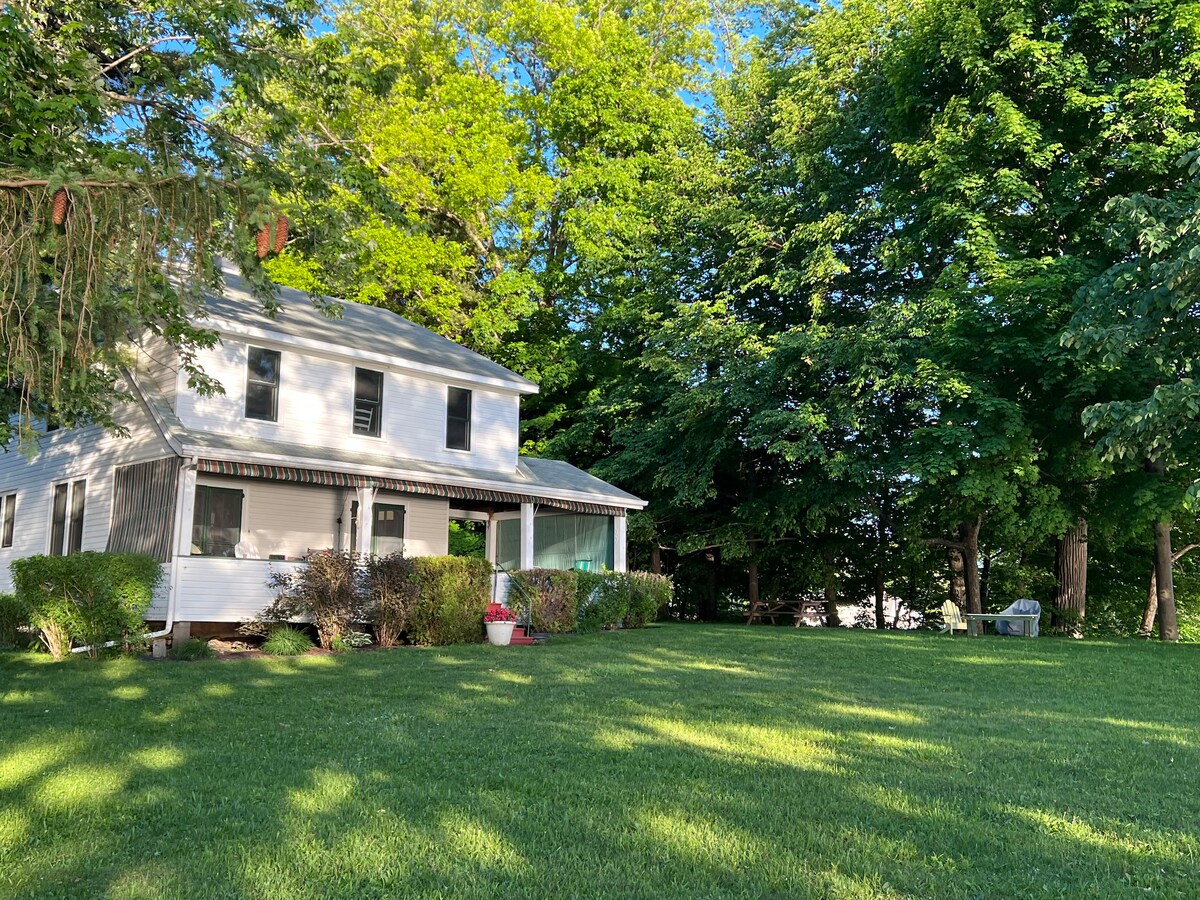 A classic two-story cottage is surrounded by lush green trees and well-maintained lawn. The front porch features large windows and is complemented by flower pots. Natural light highlights the exterior's simplicity and charm, creating a welcoming atmosphere.