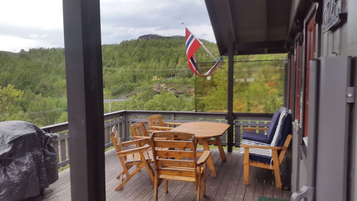 A wooden deck is featured, furnished with a large table surrounded by six chairs. A Norwegian flag is displayed proudly, with a view of lush greenery in the background. A charcoal grill is covered to the side, providing additional outdoor cooking options.