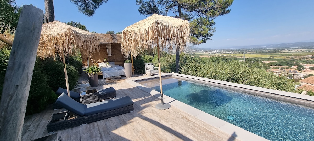 An outdoor area is revealed, featuring a private pool complemented by a deck and lush greenery. Two thatched umbrellas provide shade over lounge chairs. Scenic views of the valley extend into the horizon, framed by trees on the upper side.