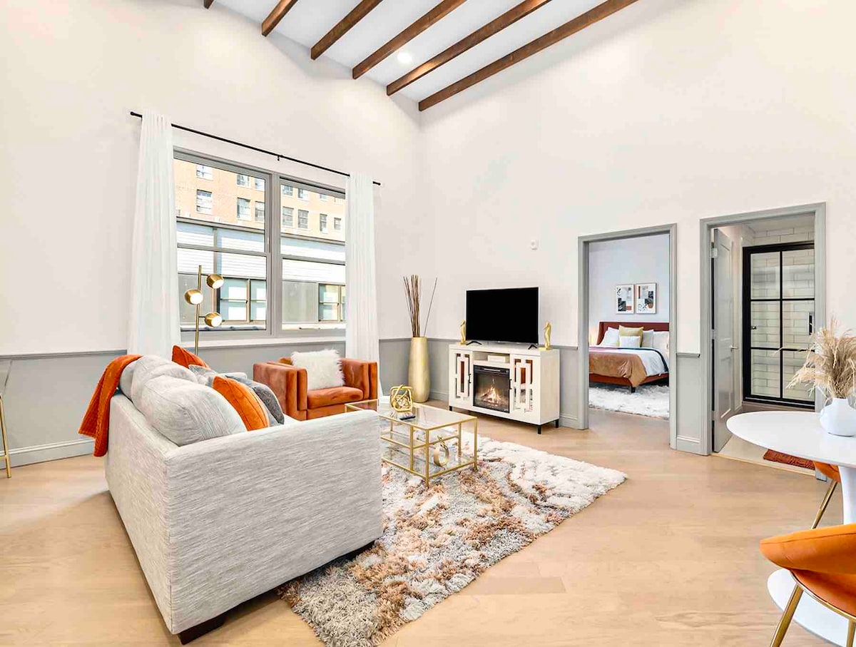 A spacious living area features high ceilings with exposed beams, complementing large windows that allow natural light to illuminate the space. Comfortable seating is arranged around a coffee table on a plush area rug. A modern entertainment unit holds a television, with a glimpse of a bedroom visible through an open door.