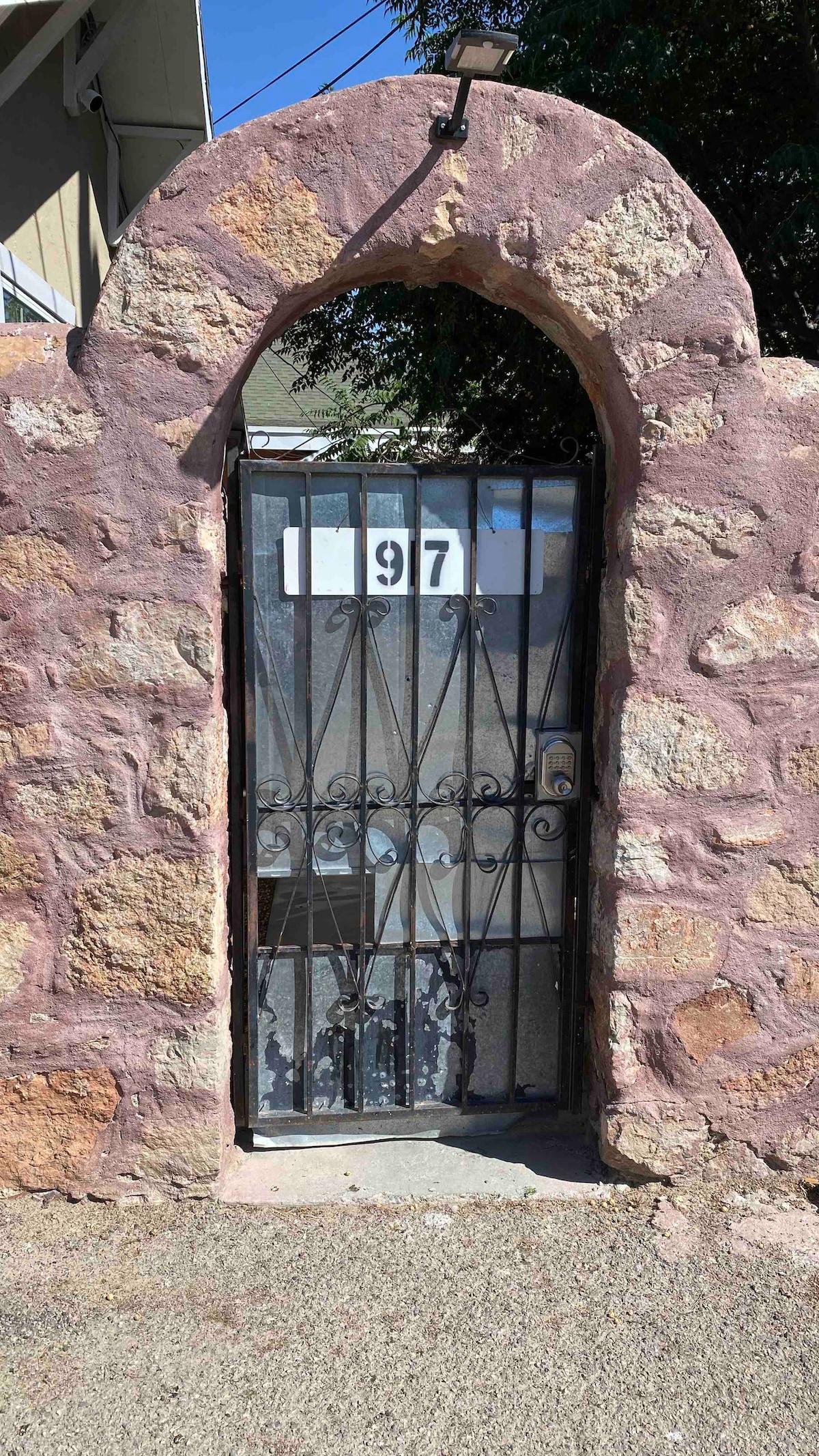 A stone archway frames a black iron gate secured with vertical bars. The gate is marked with the number '97' prominently displayed above. Sunlight casts shadows on the ground, highlighting the texture of the surrounding stone wall.