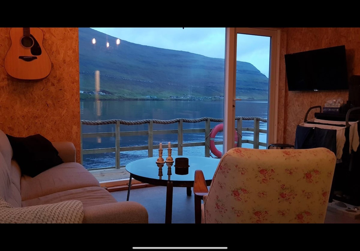 A cozy seating area features a round glass table surrounded by comfortable chairs. A guitar hangs on the wall, while a television is mounted nearby. Large windows provide expansive views of the water and surrounding landscape, enhancing the space's natural light.