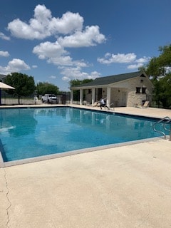 A spacious community swimming pool is surrounded by a concrete deck, featuring a diving board at one edge. A shaded area with a protective covering overlooks the pool, while green trees provide a natural backdrop. Fluffy white clouds scatter across a blue sky.