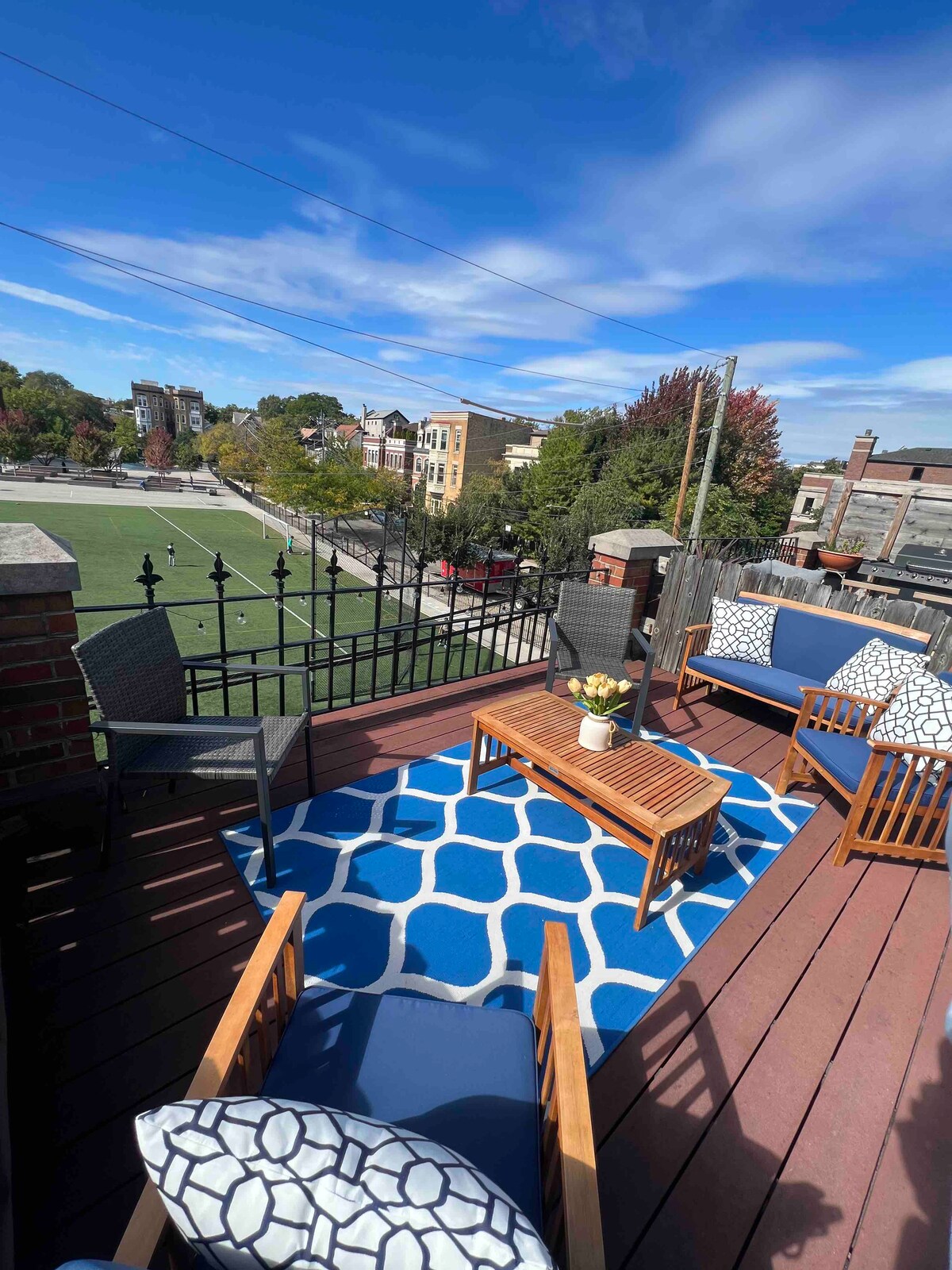 An outdoor patio space is depicted, featuring a wooden table with two chairs and a sofa set against a backdrop of urban scenery. A vibrant blue patterned rug is laid across the wooden floor. Green trees and a sports field are visible in the distance under a clear blue sky.