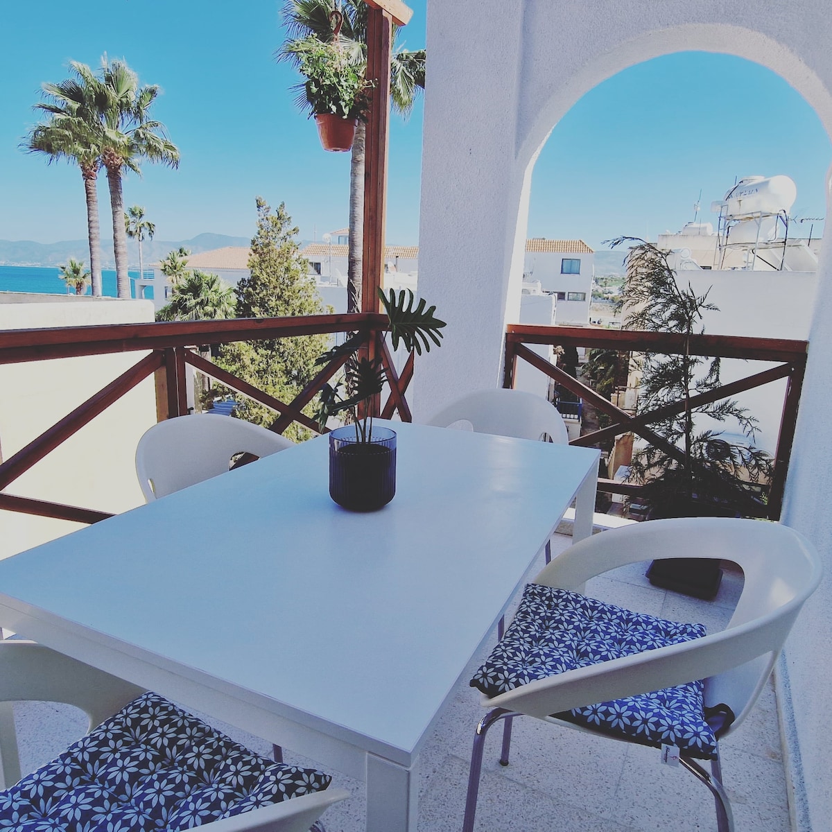 An outdoor balcony features a white table surrounded by four white chairs. Potted plants add greenery to the space, while a partial sea view is visible in the background. Clear blue skies enhance the inviting atmosphere of this outdoor area.