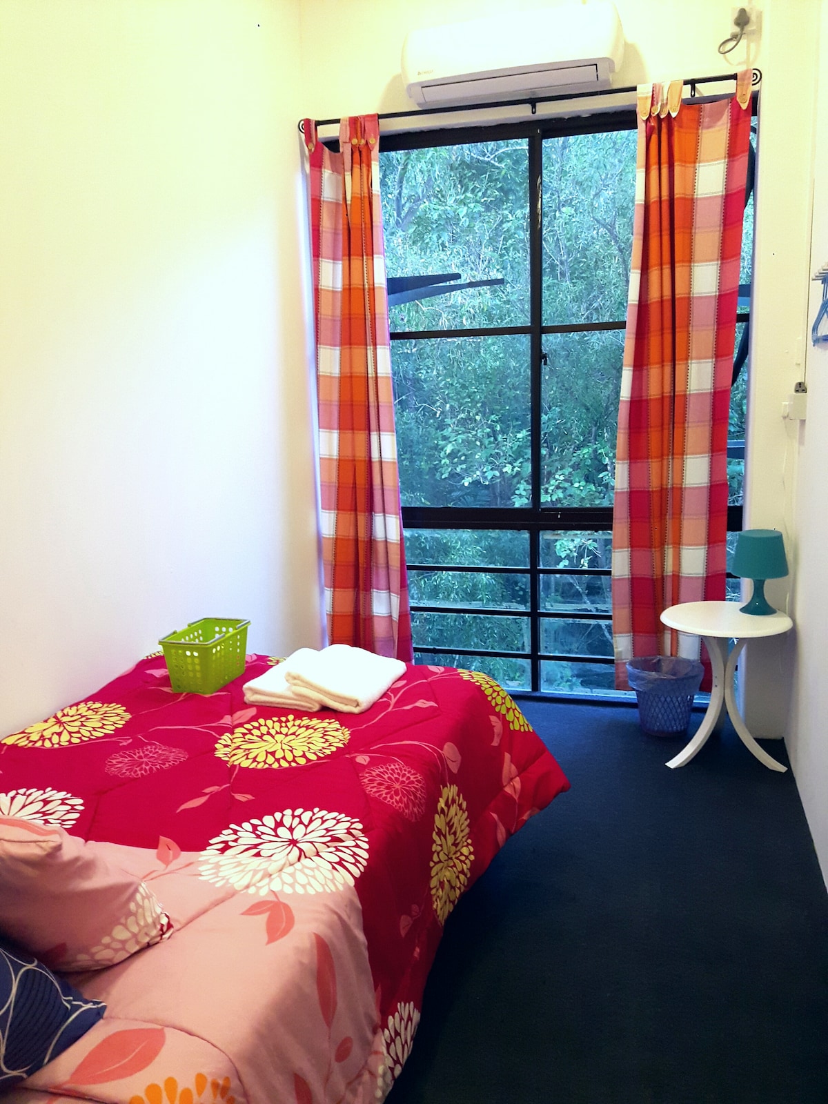 A cozy room features a single bed adorned with a floral-patterned blanket. Colorful curtains frame a large window that overlooks lush greenery. A small table and storage baskets are placed nearby, enhancing the room's functionality. Air conditioning is visible on the wall.