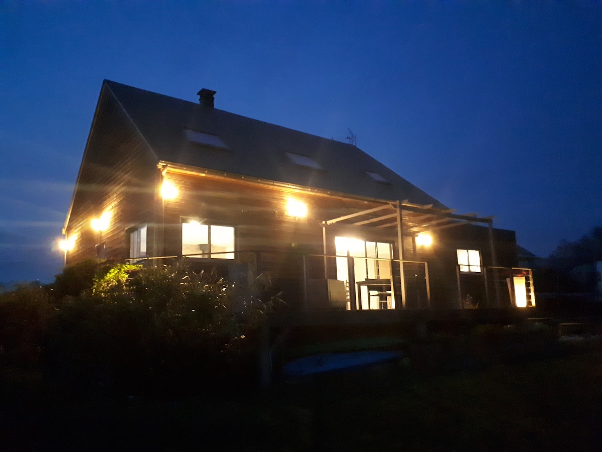 A modern wooden house is illuminated by warm lights at dusk. Large windows are visible, revealing the interior. A covered terrace extends from the building, enhancing the living space. Lush greenery surrounds the property, contributing to a tranquil setting.