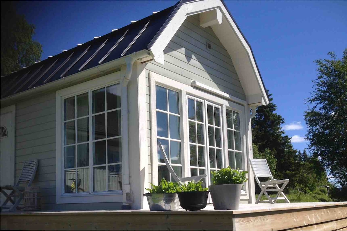 A light-colored cabin with a sloped roof features large glass windows that invite natural light. Two wooden chairs are placed on the deck, accompanied by potted plants, enhancing the outdoor space. The surrounding greenery and blue sky offer a peaceful setting.