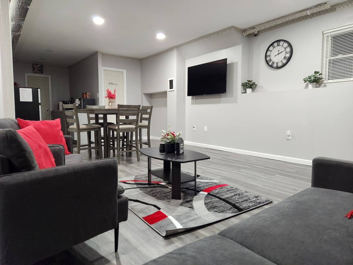 An open living area is filled with natural light and features comfortable seating arrangements. A circular coffee table sits atop a modern rug, while a dining table for six is positioned nearby. A flat-screen TV is mounted on the wall, adding to the relaxed ambiance.