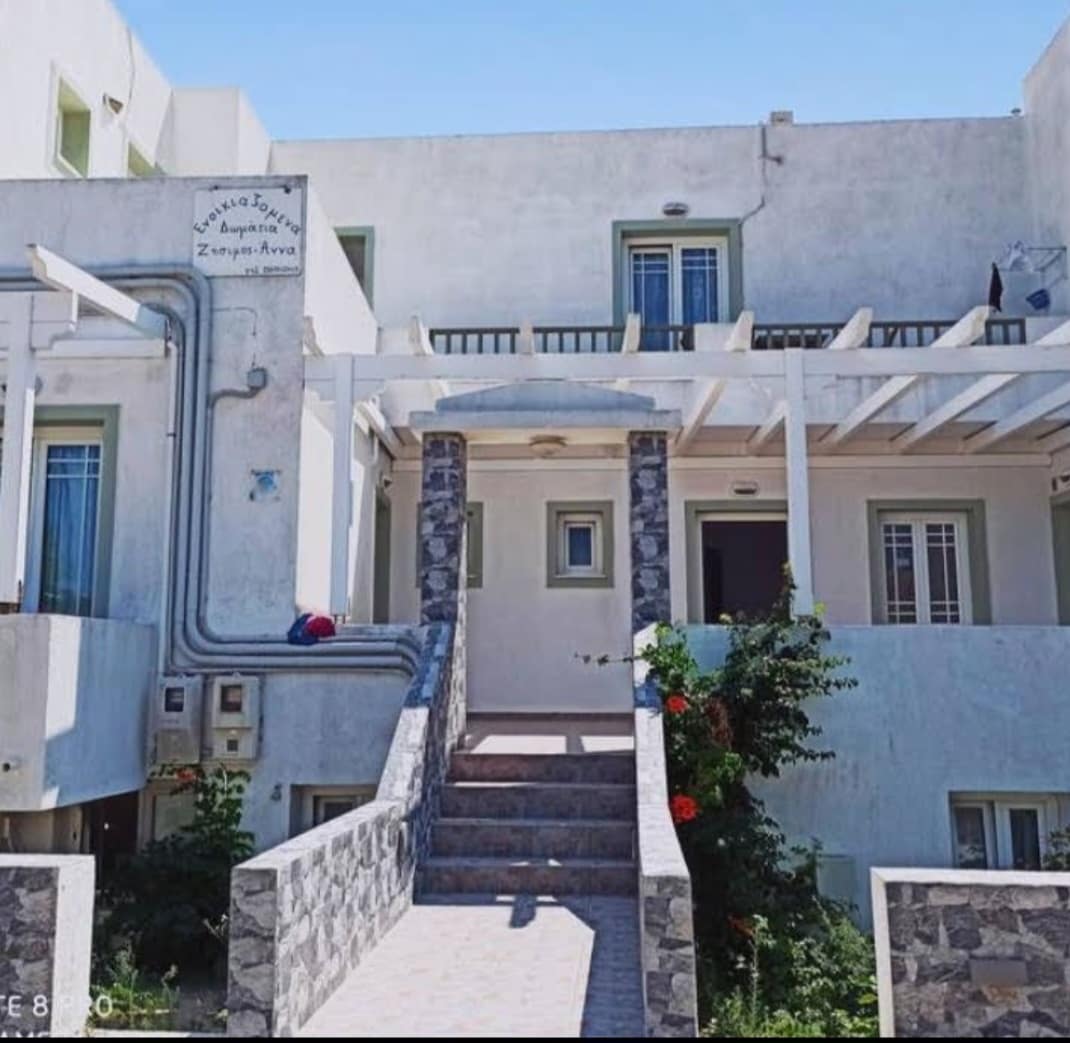 The exterior of a light-colored building is shown, featuring a staircase leading to a raised entryway. Partial shading is provided by a pergola overhead. Green plants and flowers are visible along the pathway, contributing to the serene atmosphere of the area.