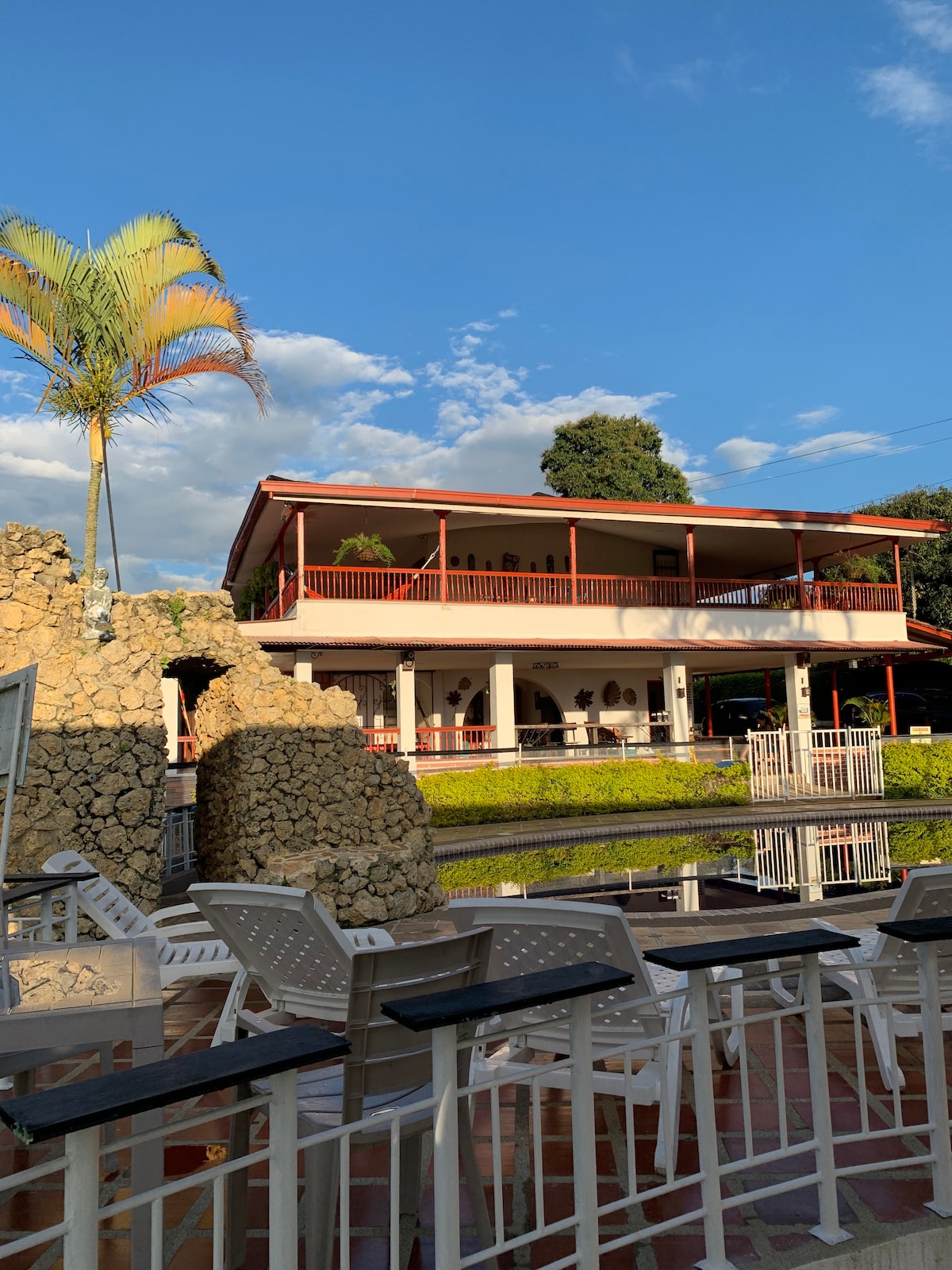 The two-story building features a traditional architecture with a red roof and white columns. Surrounding greenery complements a serene pond reflecting the structure's façade. Several outdoor seating arrangements are visible, inviting guests to relax and enjoy the peaceful surroundings.
