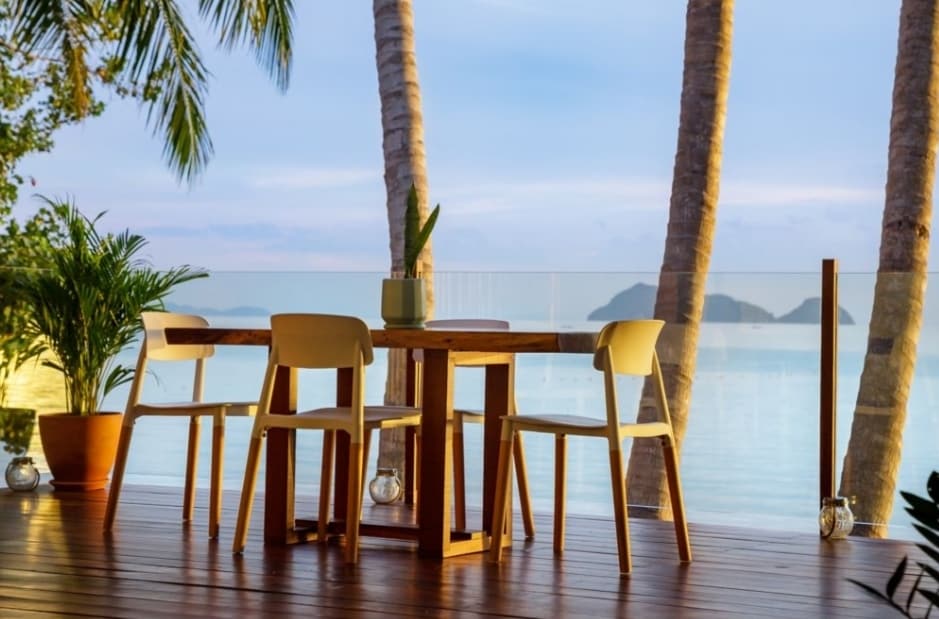 A wooden dining table with four chairs is positioned on a terrace surrounded by palm trees. The ocean view beyond the glass railing is complemented by a soft blue sky, creating a serene setting for dining or relaxation.