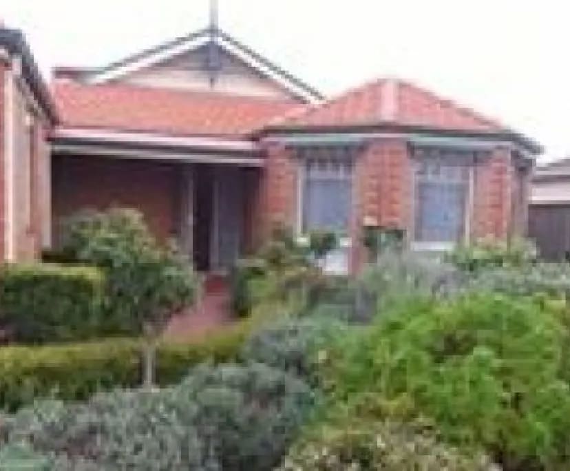 A charming house exterior is framed by well-maintained shrubs and colorful foliage. The red-tiled roof complements the brick façade, while large windows allow natural light to brighten the entrance area. A welcoming pathway leads to the main door, surrounded by greenery.