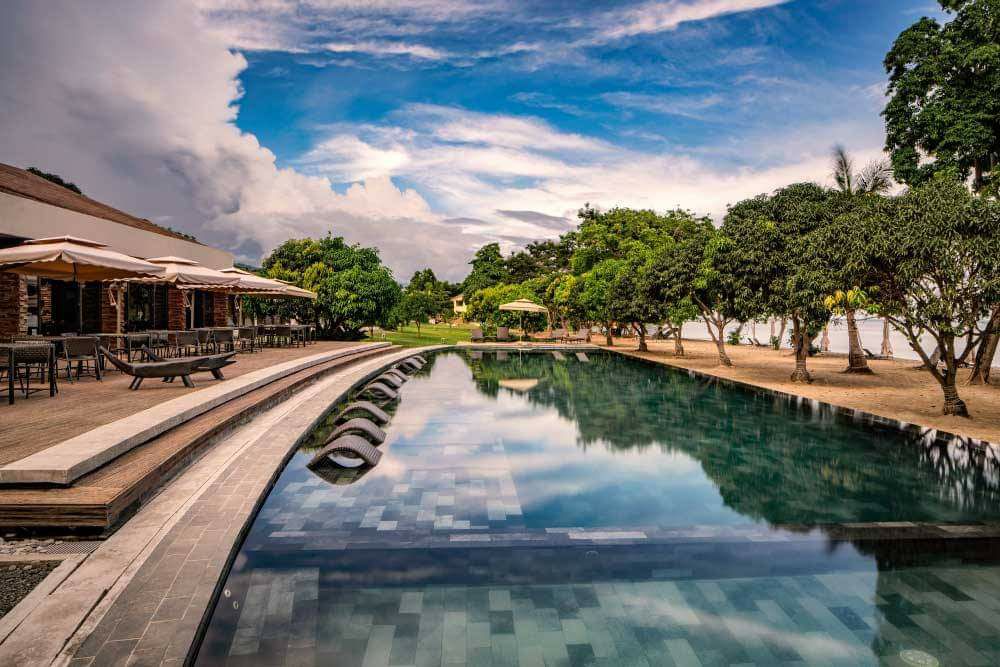An outdoor pool is surrounded by lush greenery and lounge chairs. The calm water reflects the sky and landscape, enhancing the serene ambiance. Multiple umbrellas provide shade near the pool area, and a lounging space lined with tables is visible in the background.