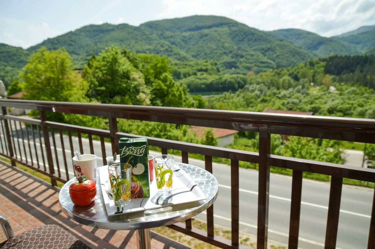 A small, round table positioned on a balcony overlooks lush green hills under a bright sky. The table is set with a tomato, glassware, and a beverage box, creating a serene spot for relaxation with nature's backdrop.