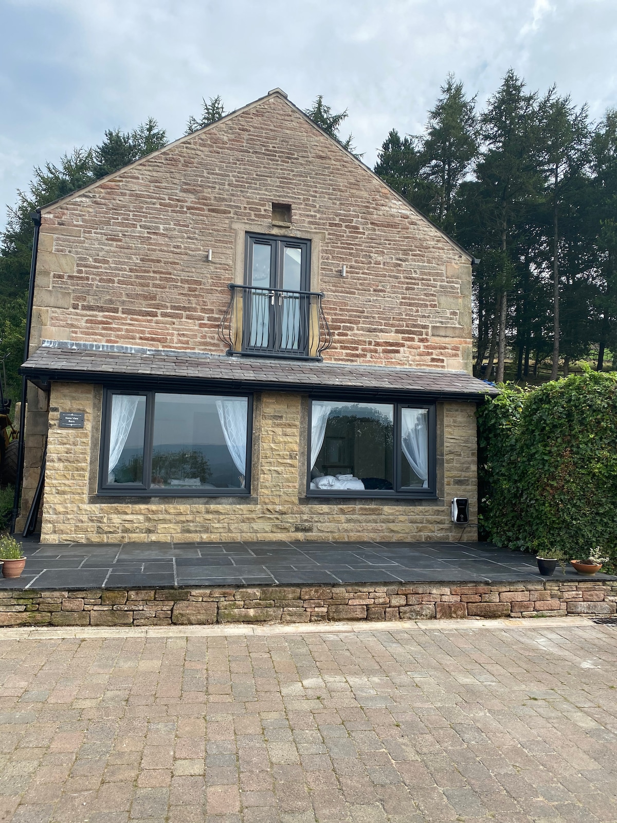 The exterior of the converted barn showcases a stone facade with large windows framing the entrance. A small patio area is visible, surrounded by greenery and potted plants. The roof features a balcony, providing additional outdoor space to enjoy the view.