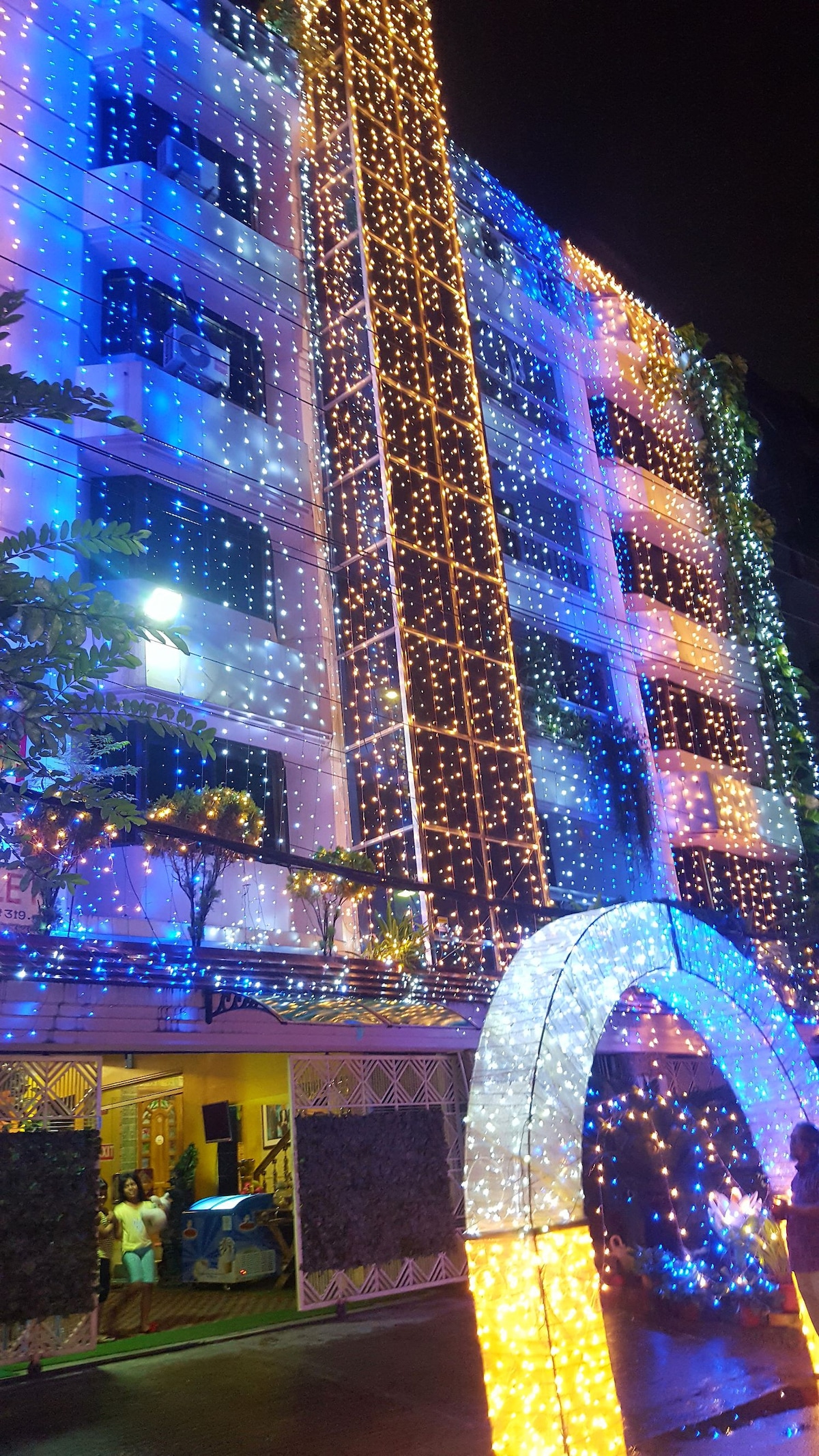 The exterior of an apartment building is illuminated by colorful string lights, creating a vibrant display. The façade features multiple levels, with greenery visible along the walls. An arched entrance is adorned with bright lights, enhancing the ambiance during the night.