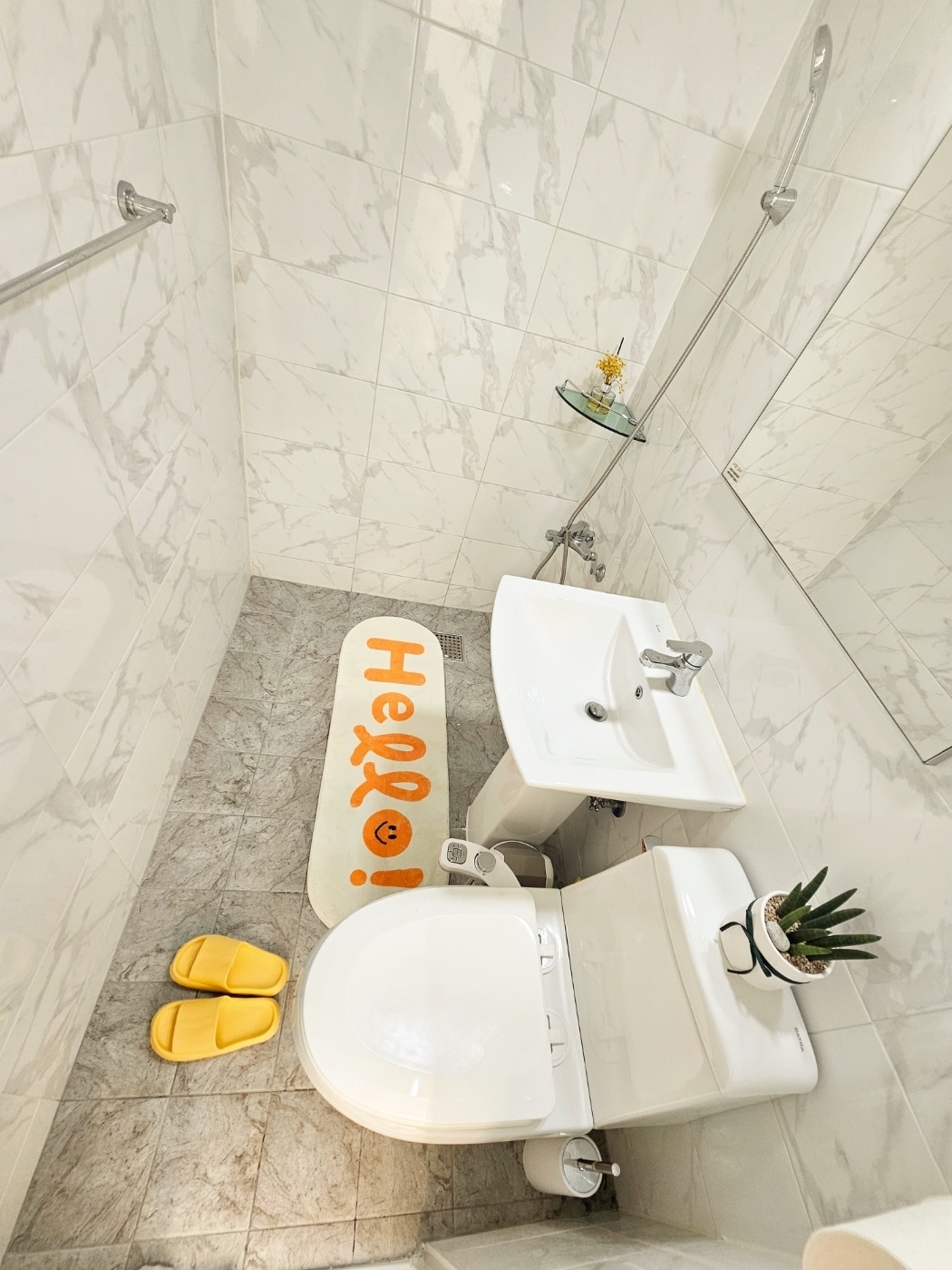 A modern bathroom features marble walls and a sleek shower area. The floor is covered with light grey tiles, and a cheerful welcome mat adds a pop of color. A small plant sits beside the compact sink, while bright yellow slippers are placed neatly on the floor.
