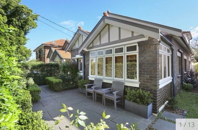 The exterior of a California bungalow is visible, showcasing a charming facade with decorative woodwork. A paved pathway leads to a landscaped area with low shrubs and plants. Two simple chairs are positioned on a small patio, offering a peaceful spot to enjoy the surroundings.