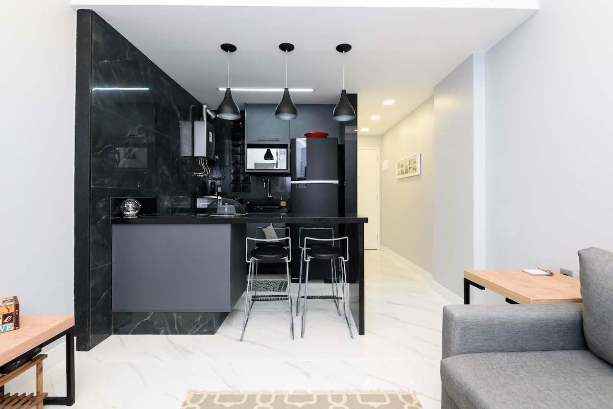 A modern kitchen area features sleek black cabinetry and marble-style walls. Three pendant lights hang above a dark countertop with bar seating. A refrigerator and stove are visible, along with a microwave. The open layout connects to a cozy living space furnished with a gray sofa.