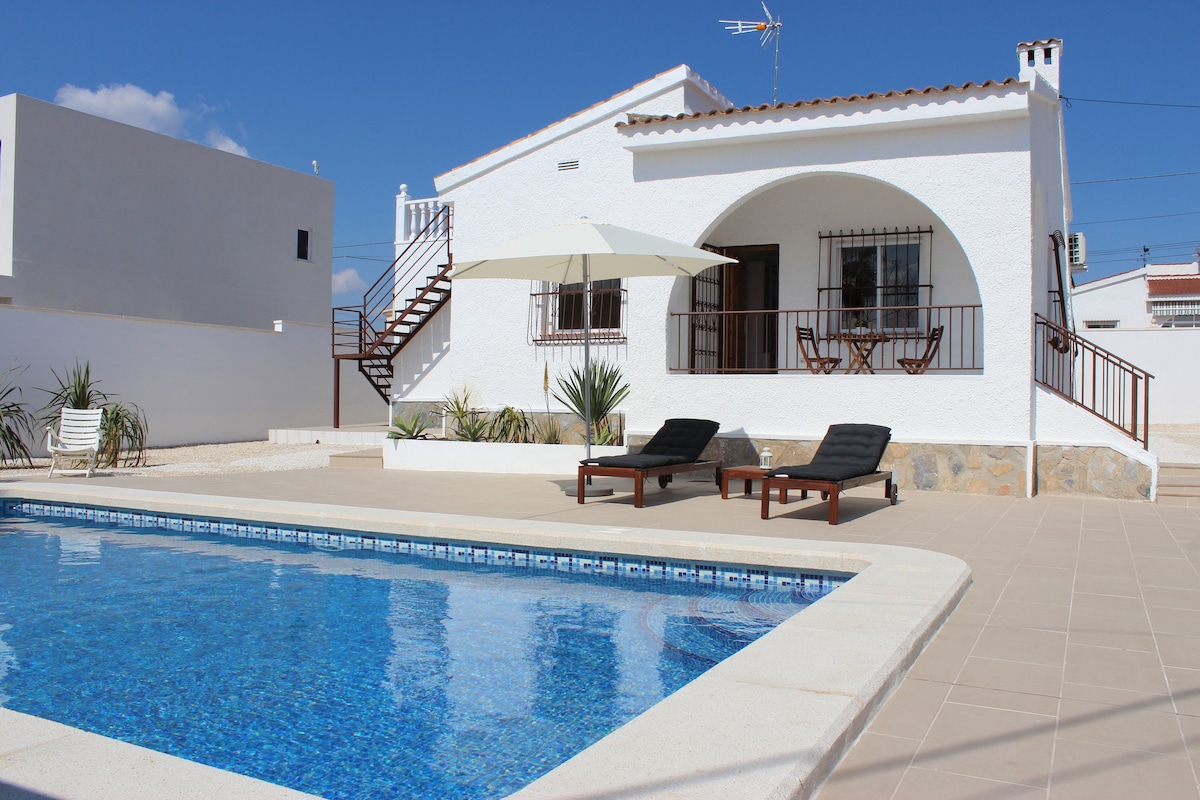 The exterior of the chalet features a private swimming pool with sun loungers positioned nearby. A shaded area is created by an umbrella, and the white building is complemented by a staircase leading to an upper level. Clear blue skies provide a bright backdrop.