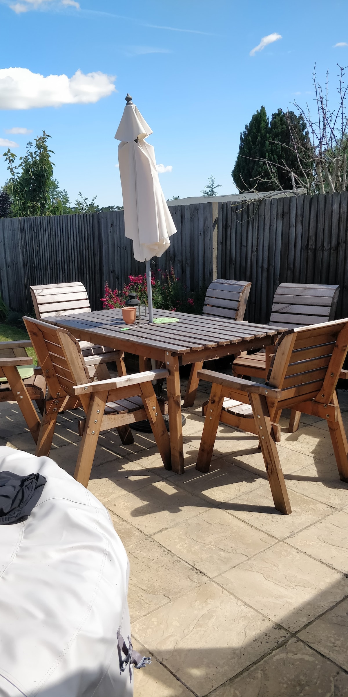A wooden patio table is surrounded by six matching chairs, all positioned on a stone surface. A large umbrella stands at the center, providing shade. Potted flowers are visible on the table, while a wooden fence and trees form the backdrop against a clear blue sky.
