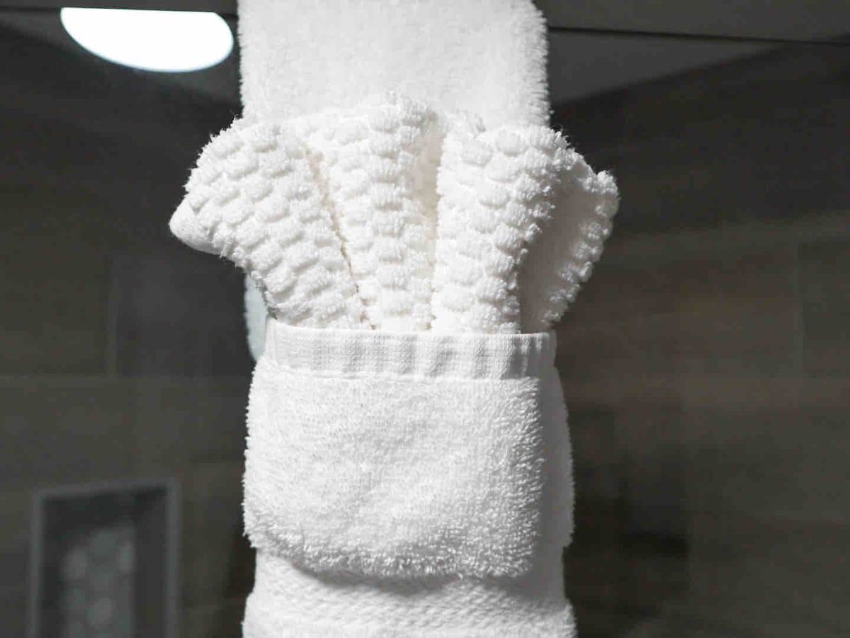 A collection of neatly arranged white towels is visible, featuring various sizes stacked together. The towels have a textured pattern, and a smaller towel is placed in a holder below the larger ones, set against a neutral background.