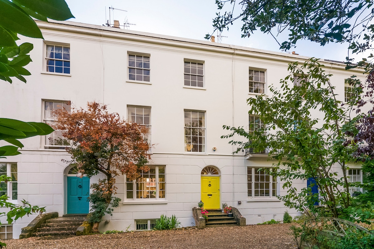 A charming townhouse exterior is showcased, featuring a white facade with large, sash windows. Two distinct front doors, one in bright yellow, stand out against the backdrop of lush greenery and colorful plants, enhancing the inviting atmosphere of the property.